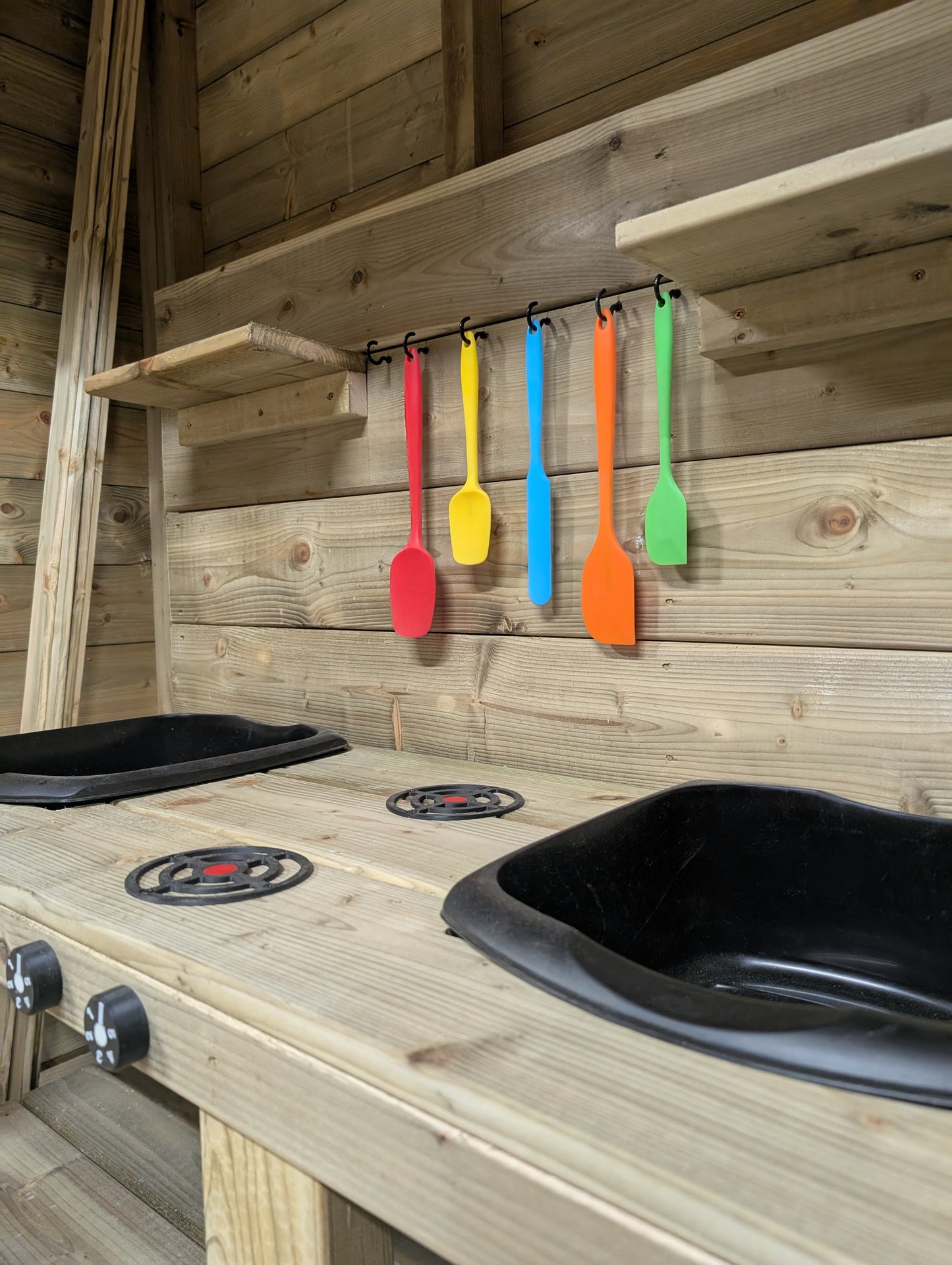 Colorful spatulas hanging on a wooden wall with a play kitchen setup.