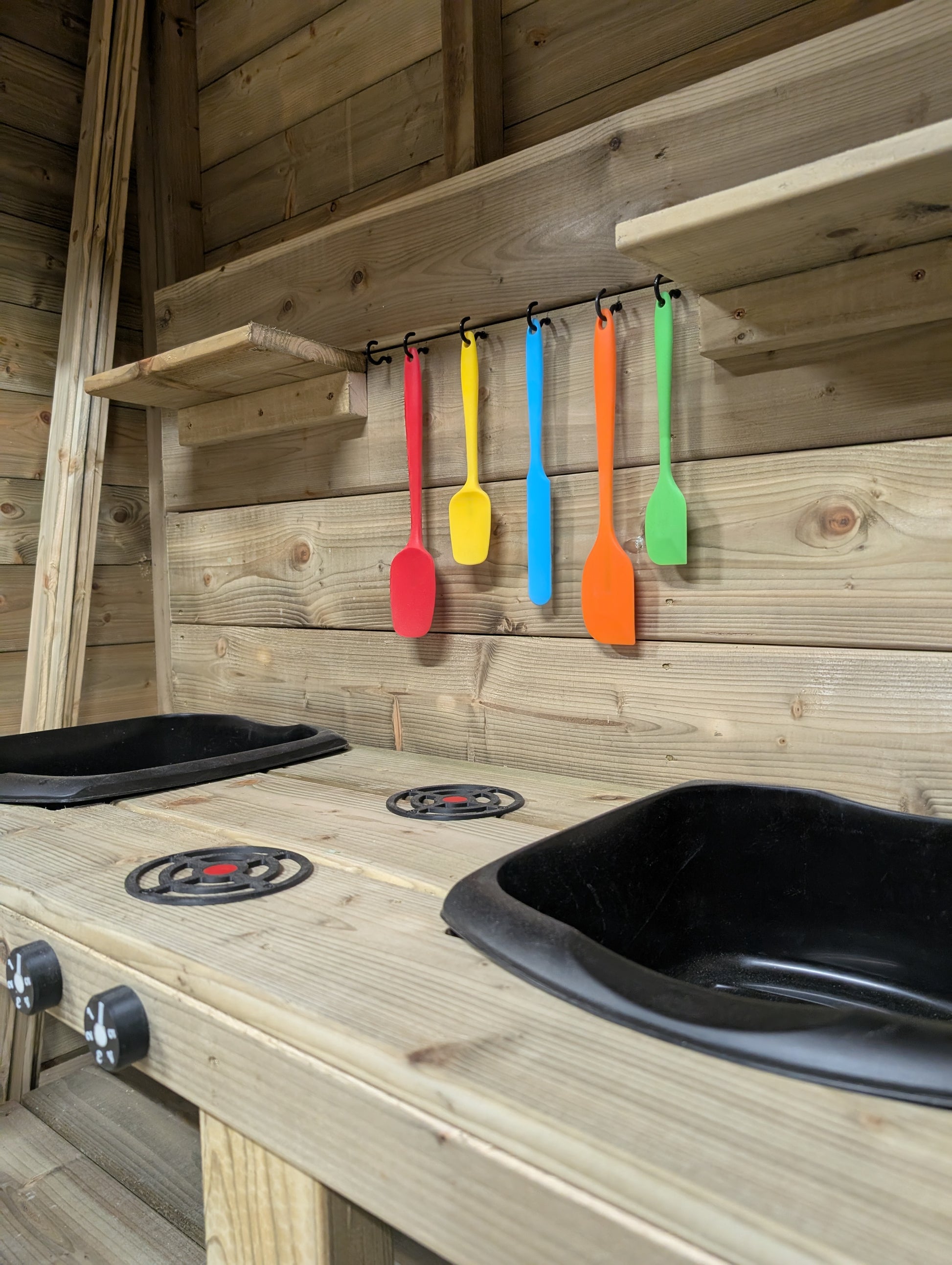 Colorful spatulas hanging on a wooden wall with a play kitchen setup.